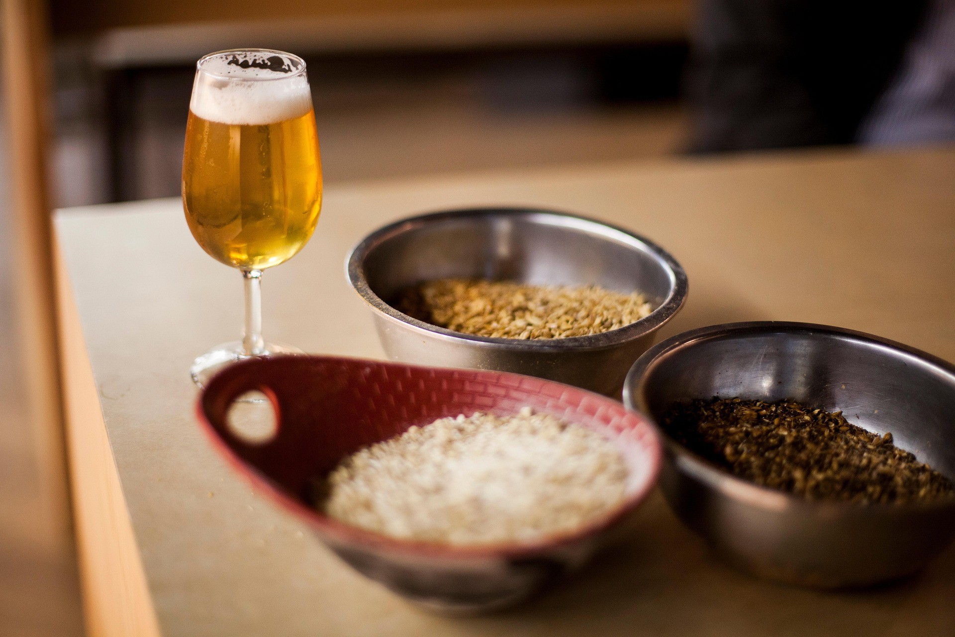 Bicchiere di birra chiara accanto a ciotole con malto d’orzo e cereali.